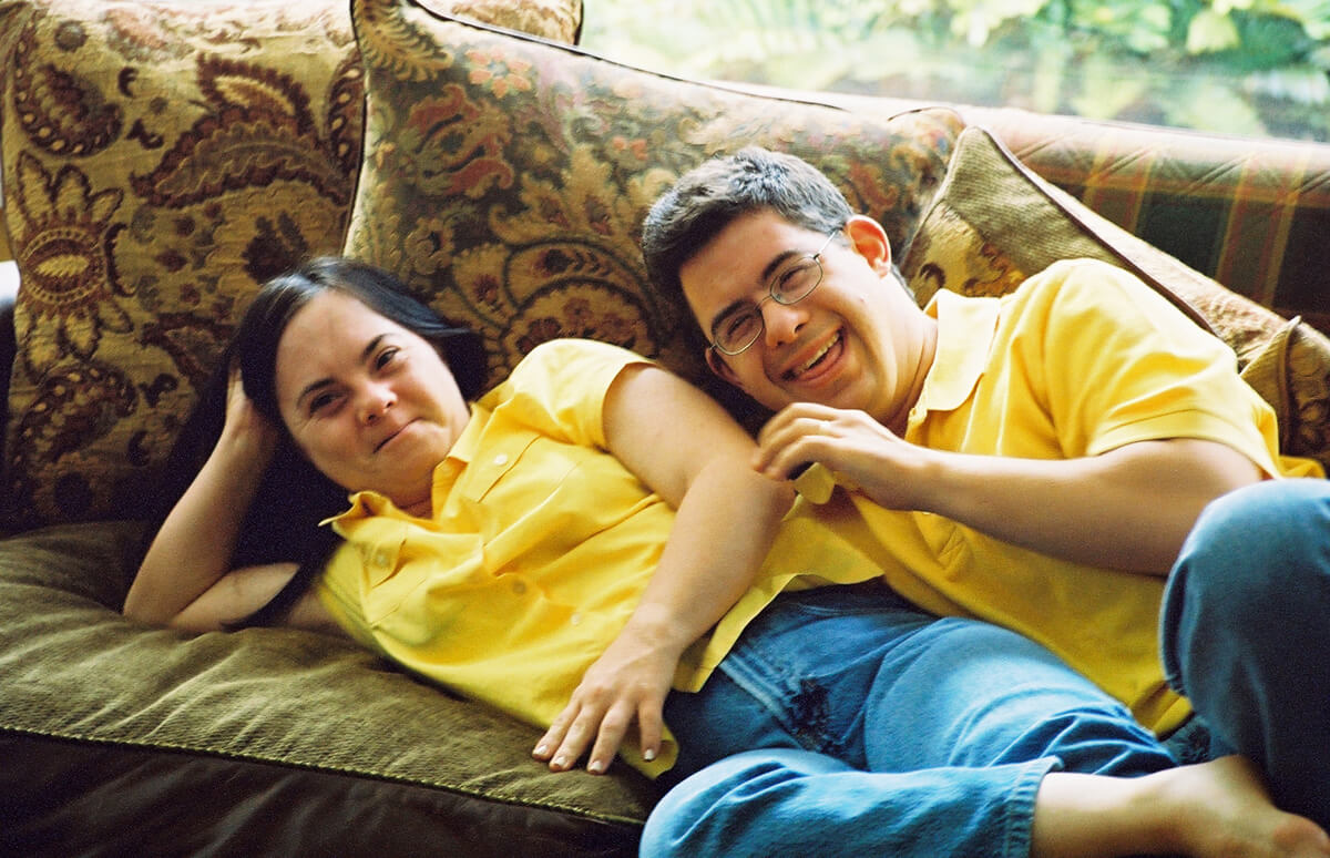 Still from Monica & David. Two people wearing yellow polo shirts and jeans are laying next to eachother on a couch with floral pillows. They are both smiling.