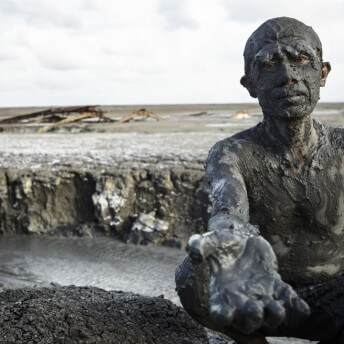 Still from Grit. A man completely covered in a thick layer of grey mud, is crouching with his right hand outstretched toward the camera. Behind him is a barren landscape of dirt and a river below.
