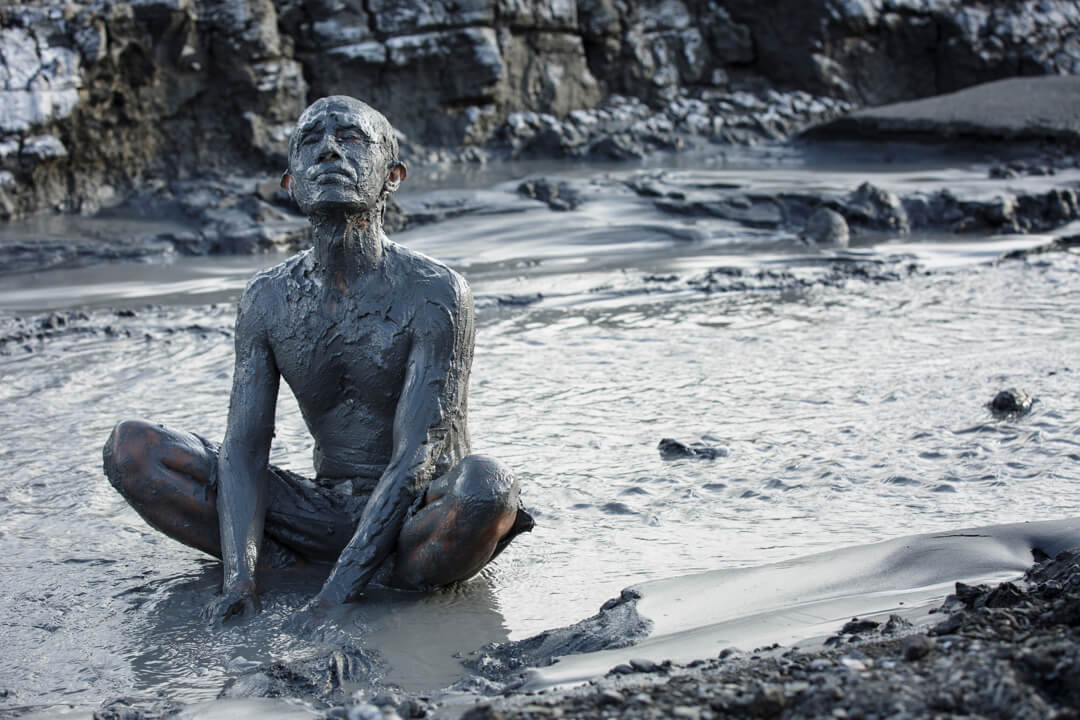 Still from Grit. A man completely covered in a thick layer of grey mud, is sitting in a flowing stream of mud.