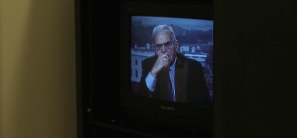 Still from My Favorite Neoconservative. A man on a television set is looking at the camera with his hand over his mouth, he is wearing glasses.