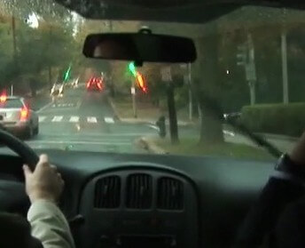 Still from My Favorite Neoconservative. A rainy dashboard view from behind a driver and passenger in a car on a street.