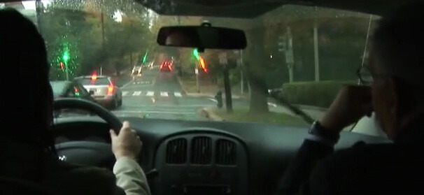 Still from My Favorite Neoconservative. A rainy dashboard view from behind a driver and passenger in a car on a street.