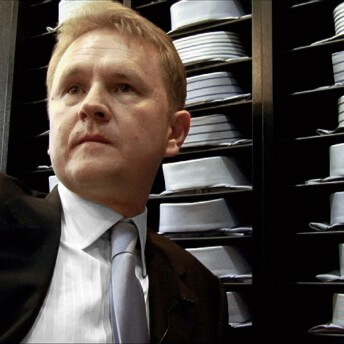 Still from My Perestroika. A man in a suit and tie stands infront rows of button up shirts folded on shelves.