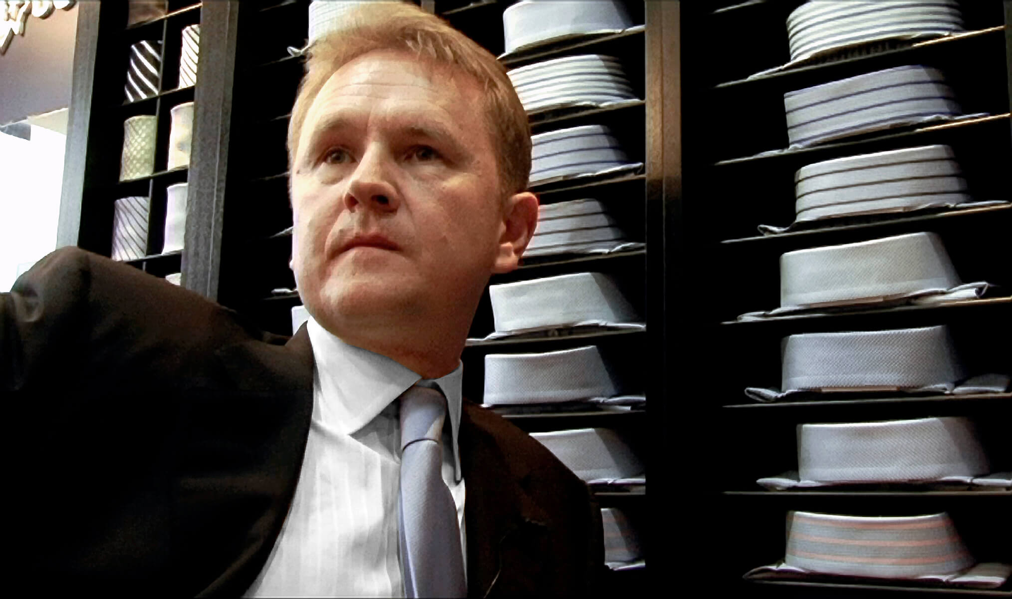 Still from My Perestroika. A man in a suit and tie stands infront rows of button up shirts folded on shelves.