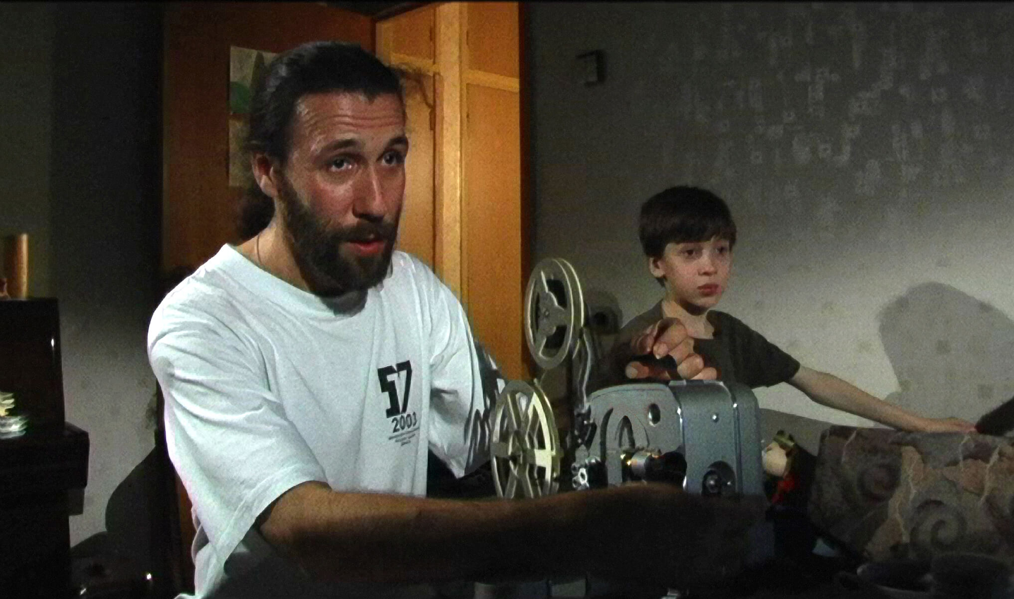 Still from My Perestroika. A man is running an older film projector. A young boy stands beside him.