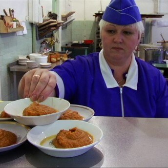 Still from My Perestroika. A woman in a kitchen, dressed in a purple uniform, balances a dish of food on top of others on display.