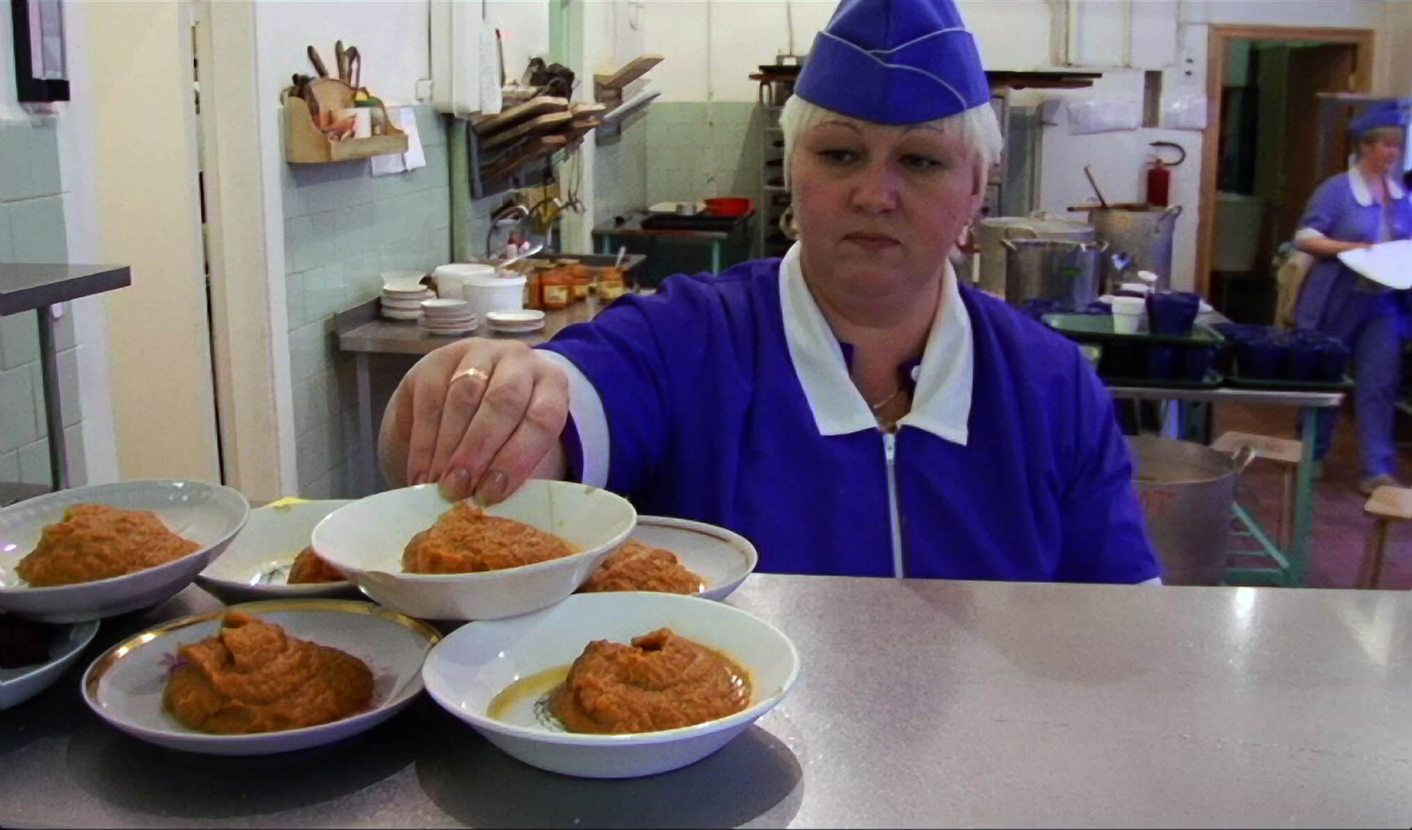 Still from My Perestroika. A woman in a kitchen, dressed in a purple uniform, balances a dish of food on top of others on display.