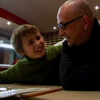 Still from My Perestroika. A man, wearing glasses, sits at a table beside a boy who is smiling and has his left arm around the man's shoulder.