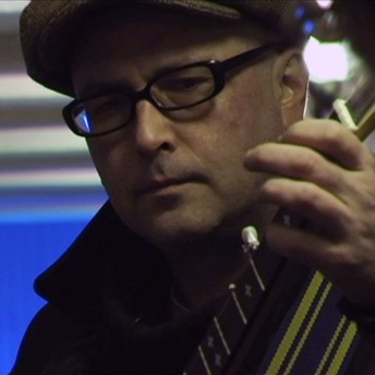 Still from My Perestroika. A man wearing glasses and a hat plays a guitar.