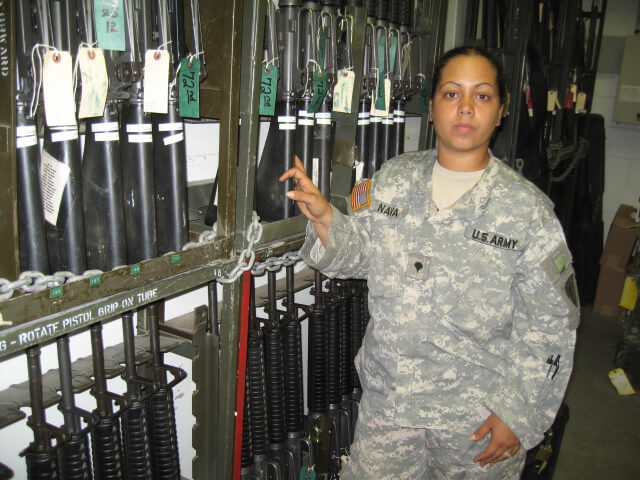Still from Lioness. A US woman soldier in uniform standing in front of a wall full of guns.