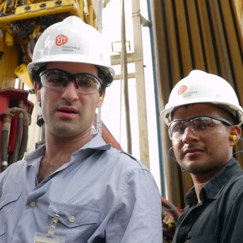 Still from Oil & Water. Two men wearing saftey glasses and helmets look at the camera. Behind them are pipes and rigging.
