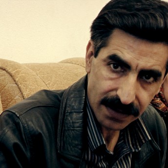 A man with a mustache and a leather jacket looks at the camera. Behind him, there is a woman wearing a scarf on her head and looking away from the camera.