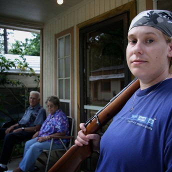 Still from Lioness. In the foreground there is a woman in a blue t-shirt and a head bandana holding a gun that rests on her shoulder, she looks directly at the camera. In the background there is an elderly couple sitting on chairs.