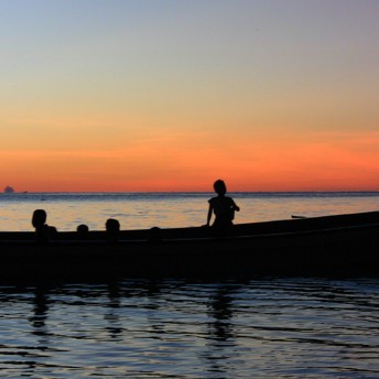 A photo of four children sitting in a boat on the water. It is sunset, the water is dark blue and the sky features a vibrant sunset of yellows, reds, and oranges. Only the silhouette of the children on the boat is visible.