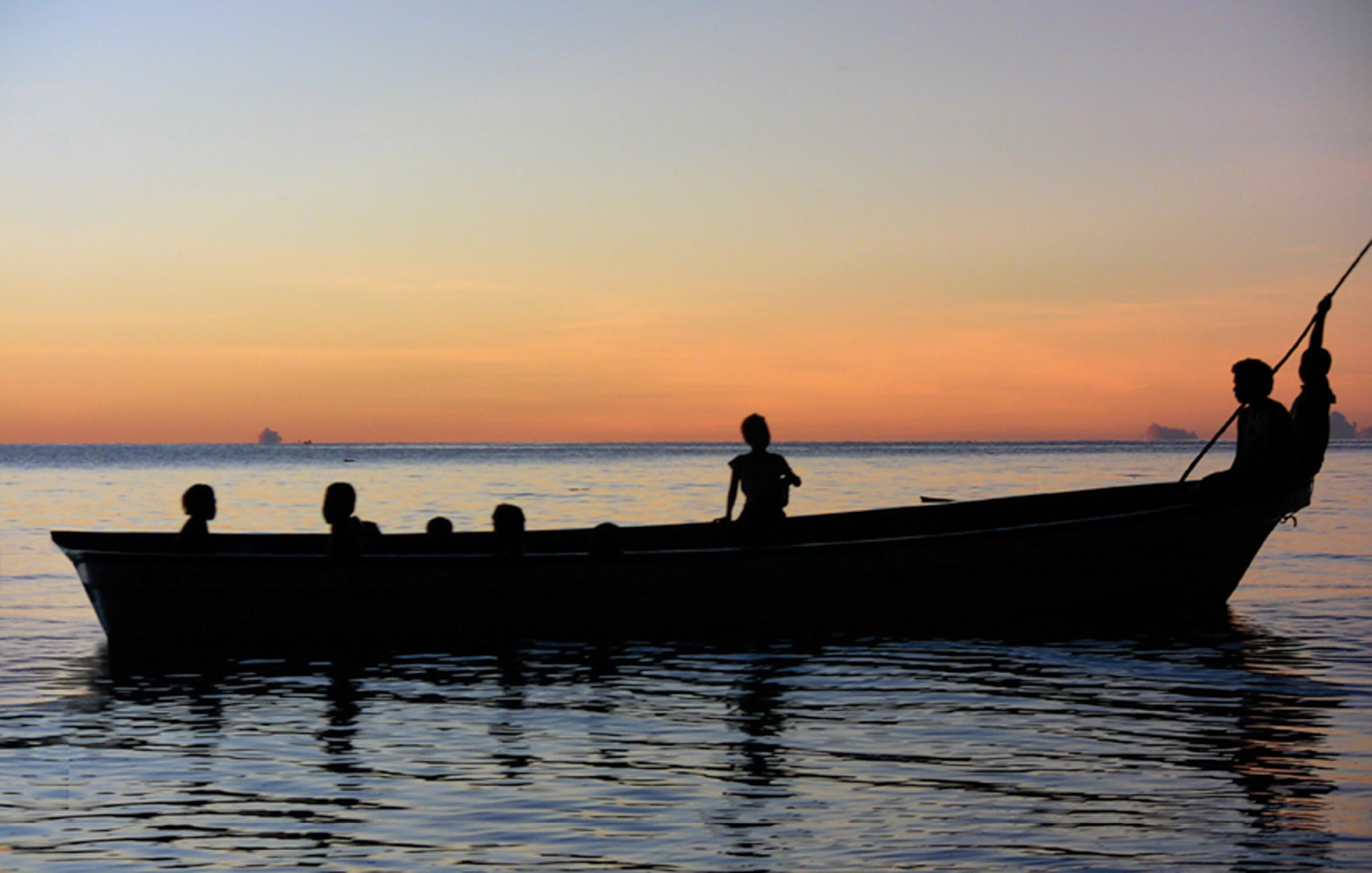 A photo of four children sitting in a boat on the water. It is sunset, the water is dark blue and the sky features a vibrant sunset of yellows, reds, and oranges. Only the silhouette of the children on the boat is visible.