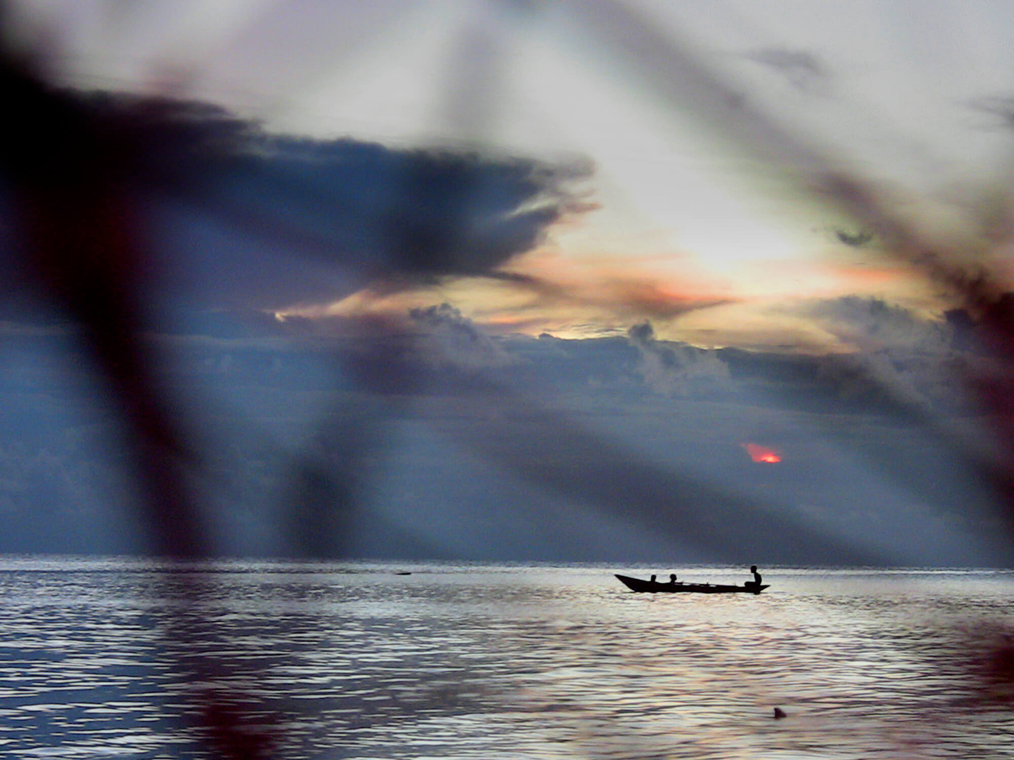 A long shot of three people seated in a small motorboat on the water at dusk. There are branches in the foreground, close to the camera and out of focus.