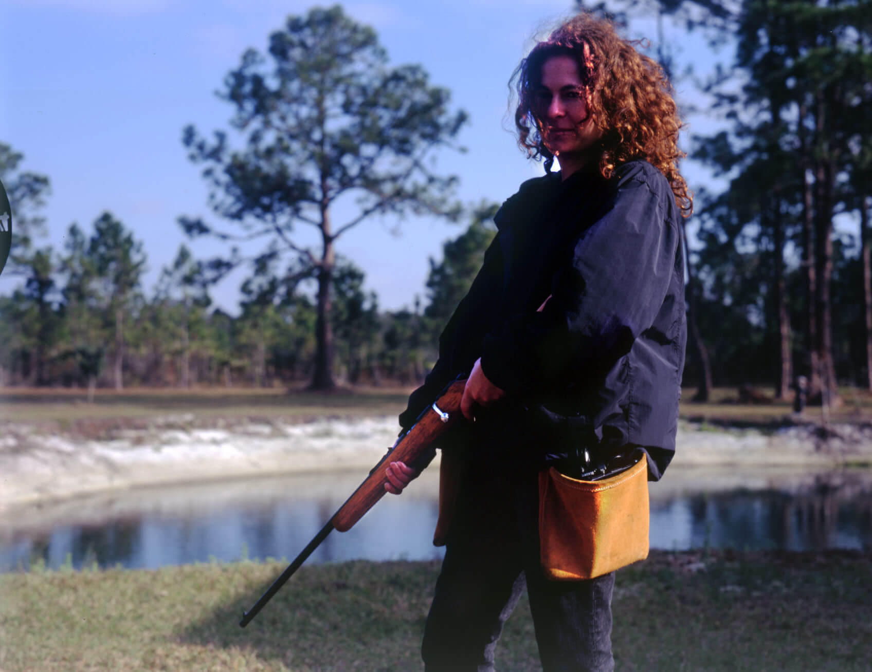 Julie Kahn stands slighty far away from the camera, she is standing on a patch of grass in front of a pond. She is turned to the side away from the camera with her head turned to the left to face the camera. She has slightly longer than shoulder length hair that is brown and curly. She is wearing jeans and a blue jacket with a yellow bag hanging at her left hip. She is holding a shotgun pointed down towards the ground.