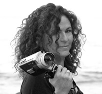 Julie Kahn turns her head to the right to look directly into the camera. Portrait in black and white. She holds a camcorder in her right hand.