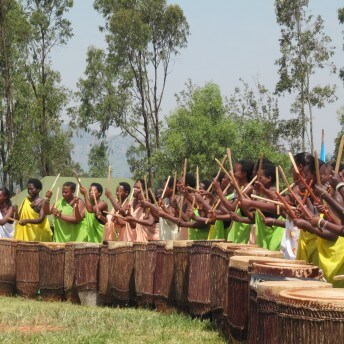 A still from Sweet Dreams. A line of women stand in the middle of a field, each in front of a waist-high drum. They are holding drumsticks and their arms are raised above their heads, mid strike. They all wear one shoulder wraps in various shades of greens, pinks, and whites.