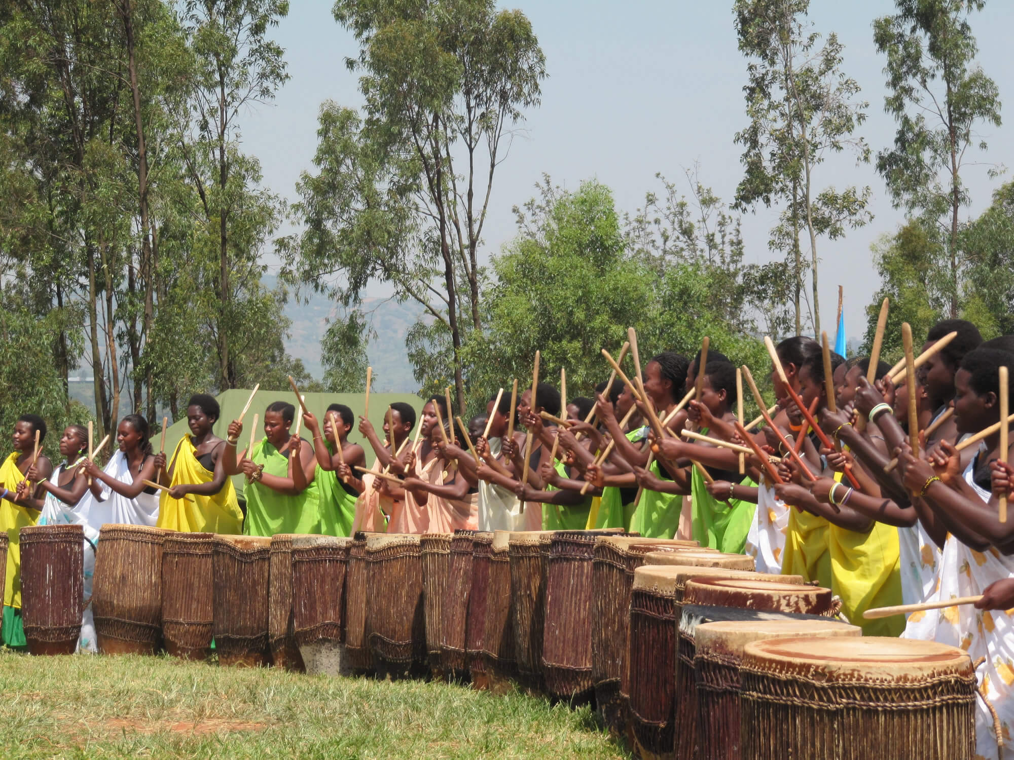 A still from Sweet Dreams. A line of women stand in the middle of a field, each in front of a waist-high drum. They are holding drumsticks and their arms are raised above their heads, mid strike. They all wear one shoulder wraps in various shades of greens, pinks, and whites.