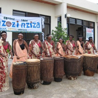 Still from Sweet Dreams. A line of eight women faces the camera. They are smiling and playing stomach-height wooden drums. They stand shoulder to shoulder in front of a white banner that reads, "Coming Soon Zozi nziza: cream, coffee, dreams".