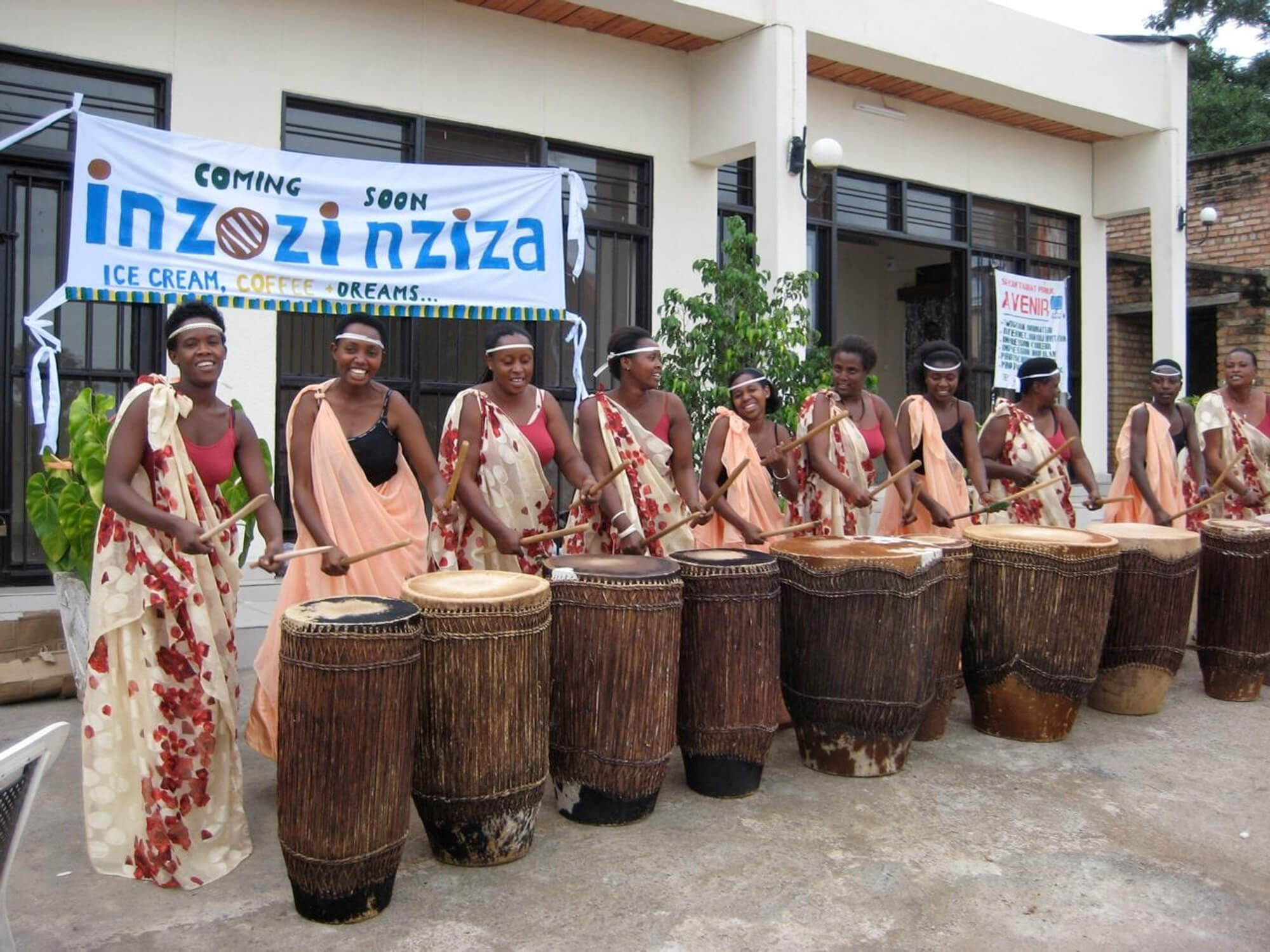 Still from Sweet Dreams. A line of eight women faces the camera. They are smiling and playing stomach-height wooden drums. They stand shoulder to shoulder in front of a white banner that reads, "Coming Soon Zozi nziza: cream, coffee, dreams".