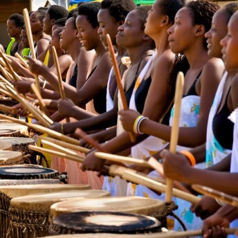 A line of women holding drumsticks, arms bent, mid drum-strike over stomach-height wooden drums stretches from the foreground into the background. All of the women are wearing black tank tops and a patterned one-shoulder wrap overtop.