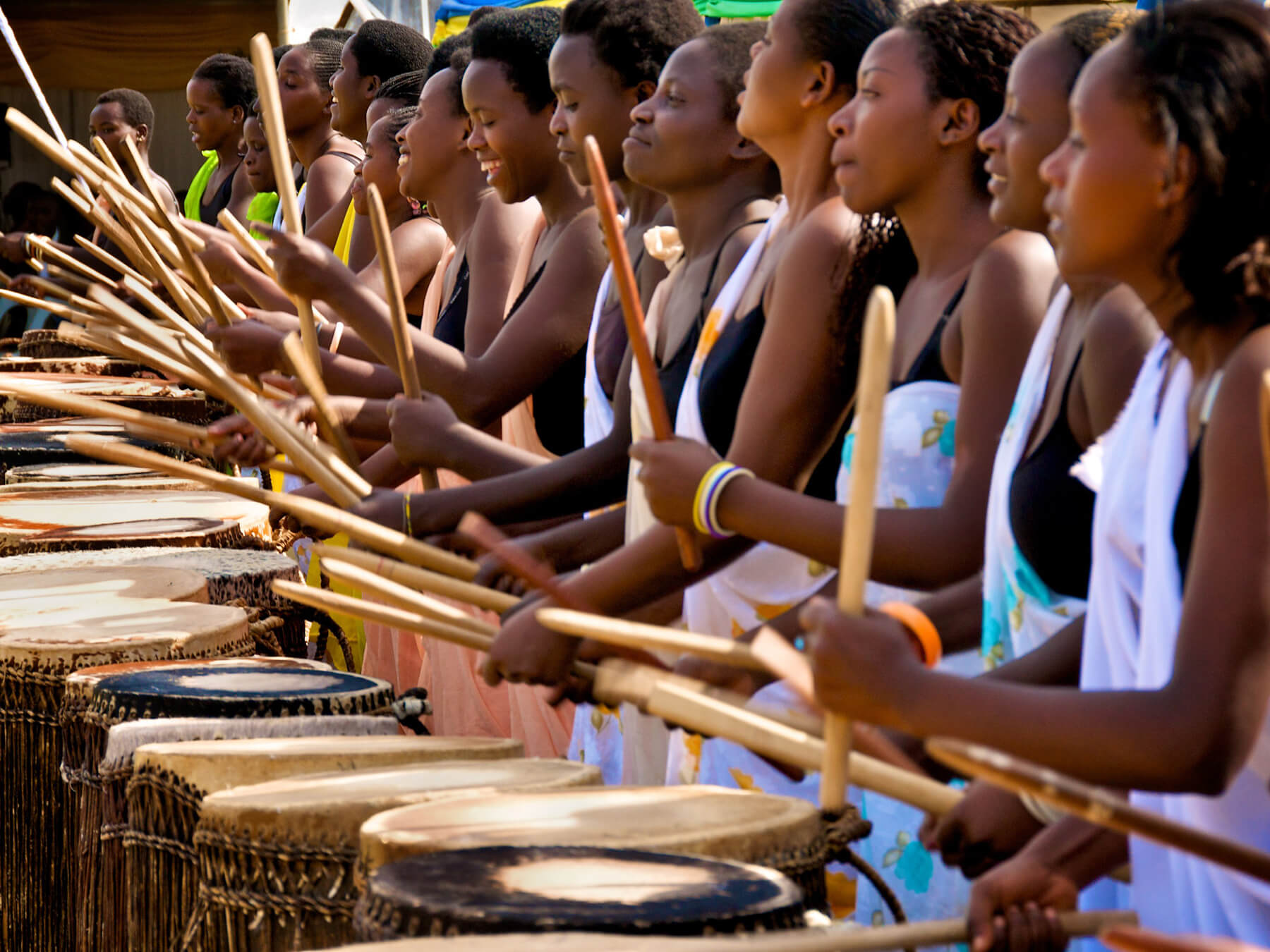 A line of women holding drumsticks, arms bent, mid drum-strike over stomach-height wooden drums stretches from the foreground into the background. All of the women are wearing black tank tops and a patterned one-shoulder wrap overtop.