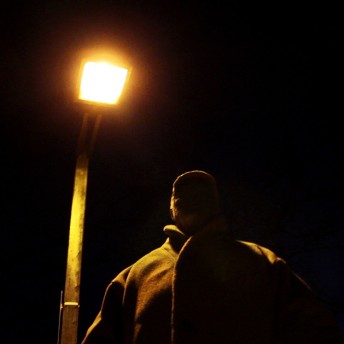 Still from (T)ERROR. A man standing under a streetlamp looks down at the camera. It is night time, he is wearing a baseball cap and a jacket. None of his features are visible.