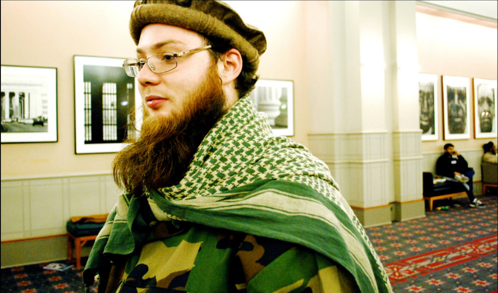 Still from (T)ERROR. A man with a beard faces to the right. He has a large beard and is wearing all green.