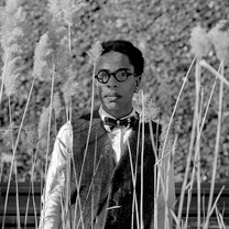 Lyric R. Cabral stands in a field of tall grass and looks directly at the Camera. Portrait in black and white.