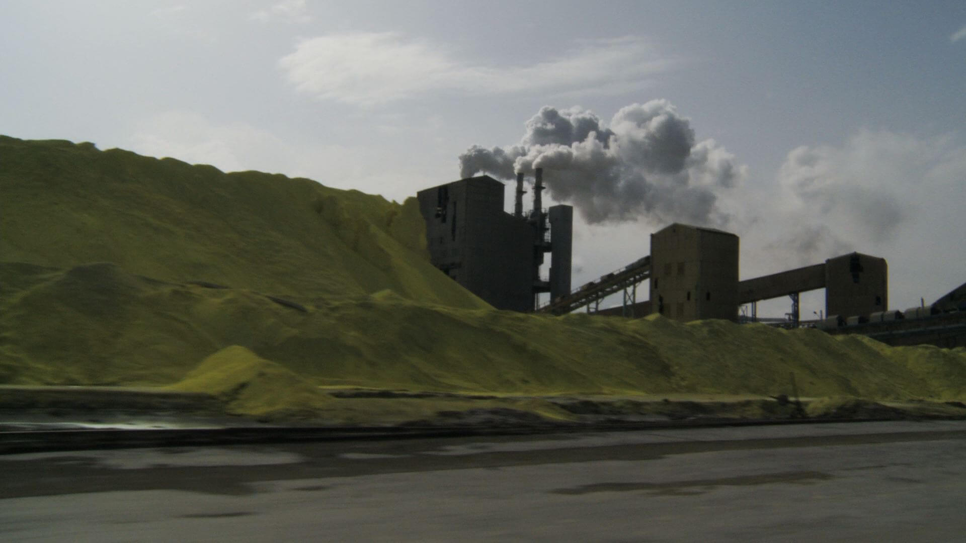 A still from The Factory and Me. A factory is nestled next to a series of green hills on the opposite side of a river. White smoke billows from the pylons.