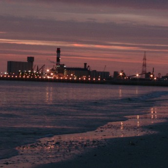 A still from The Factory and Me. The sun sets on a beach. There is a factory on the shoreline, off in the distance.