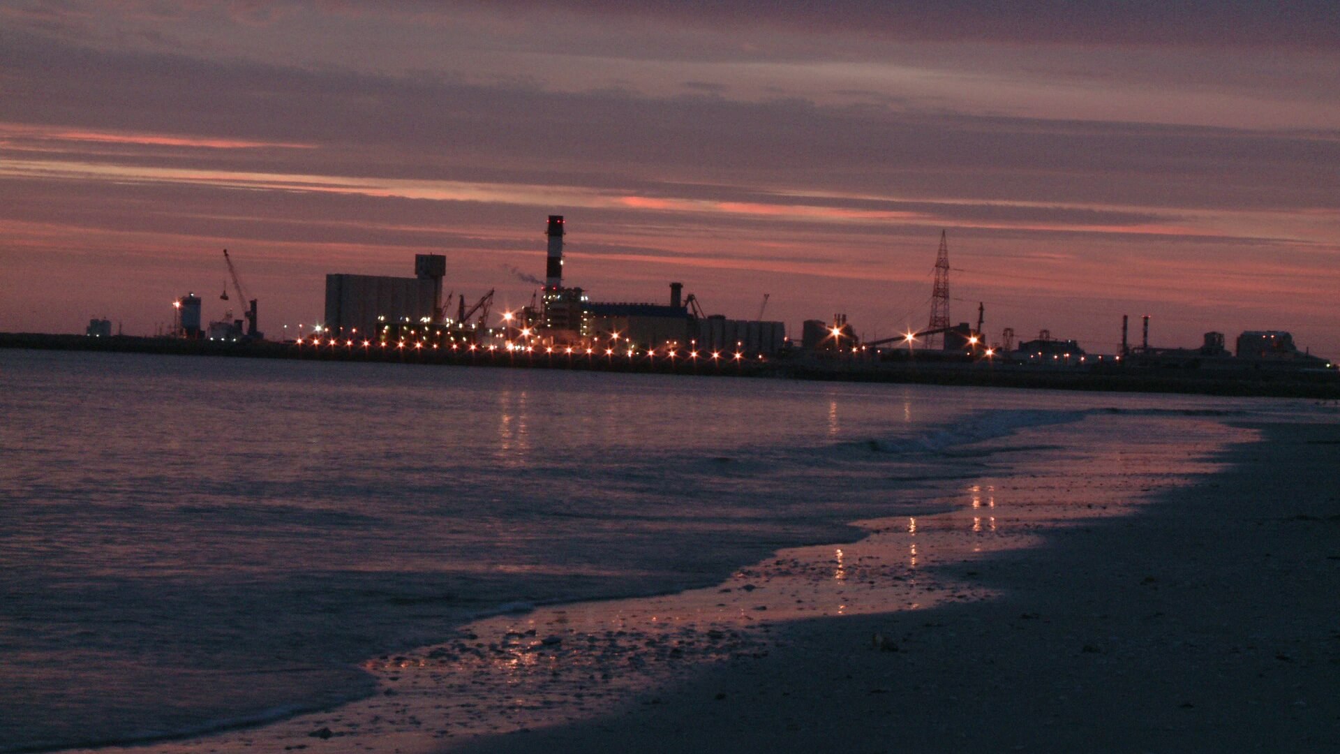 A still from The Factory and Me. The sun sets on a beach. There is a factory on the shoreline, off in the distance.