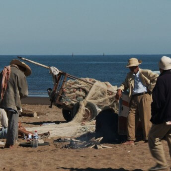 A still from The Factory and Me. Three people stand in front of a large cart filled with plastic bottles and a net. They are on the beach in the sun.