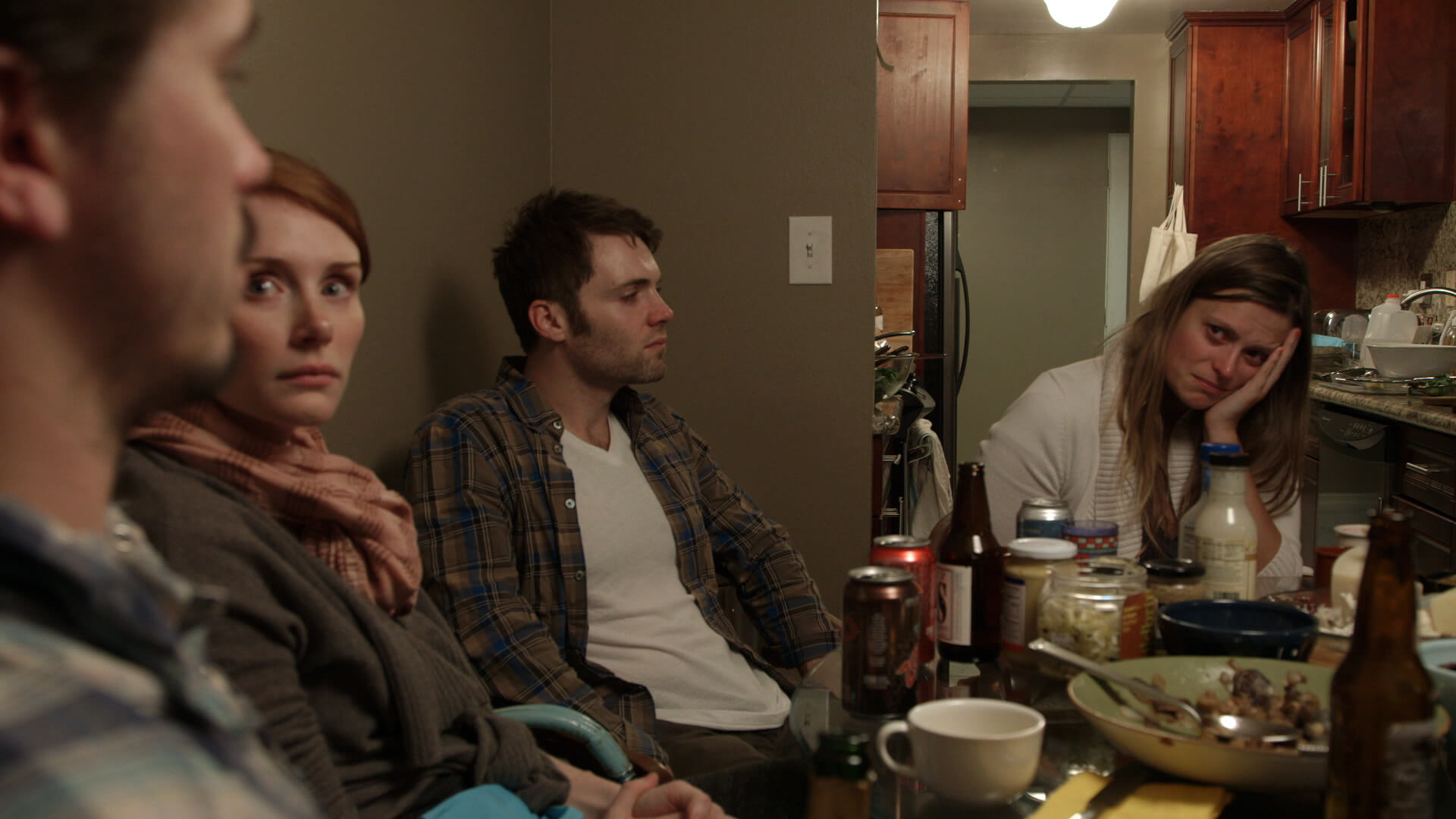 Still from The Lion's Mouth Opens. Close up shot of four people sitting around a table. On the table rests and assortment of dishware, jars, and cans.