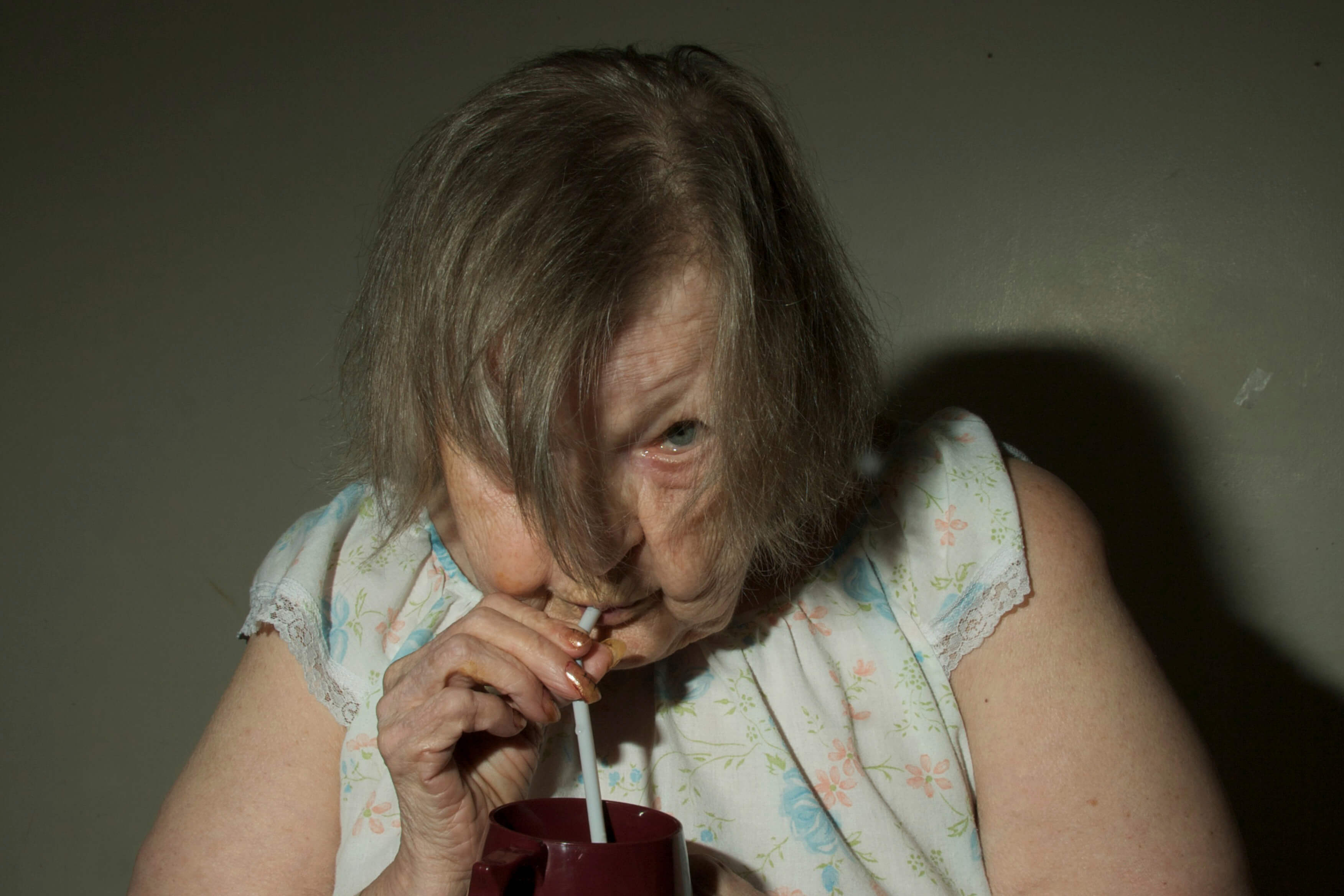 Still from The Patron Saints. A person stands in front of the camera wearing a sleeveless nightgown. They hold a straw to their mouth and peer up at the camera from between their hair which falls down over each side of their face.
