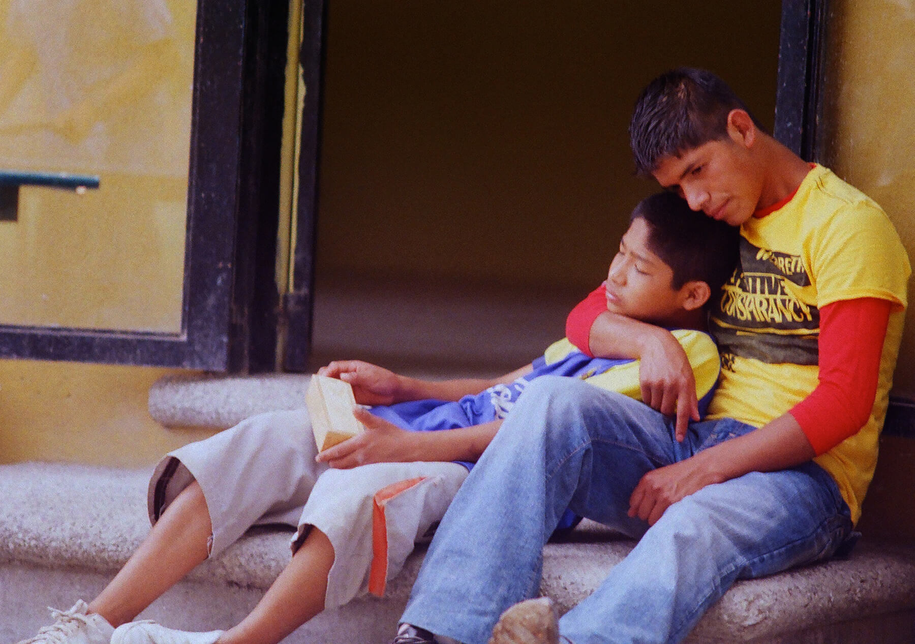 Still from Visitor's Day. A young man sits with a young boy on the ground outside, leaning on each other. The older one with an arm around the young boy.