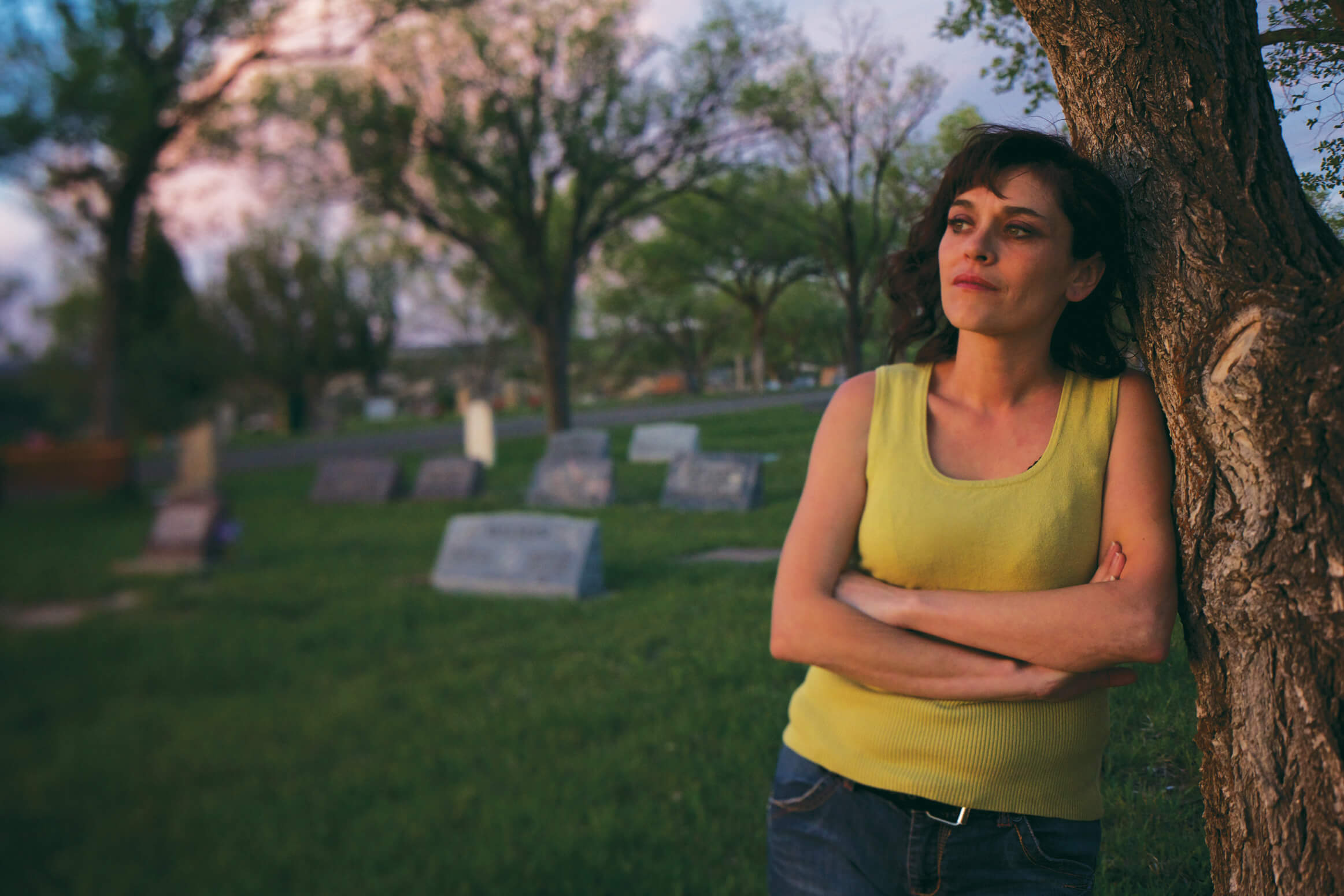 Still from Uranium Drive-In. A woman in a yellow tanktop stands in a cemetery, leaning against a tree.