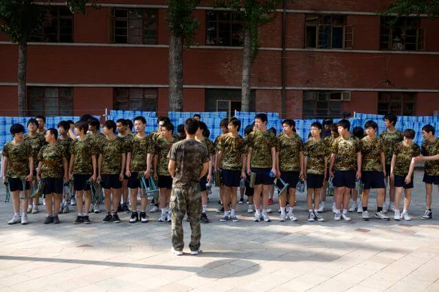 Still from Web Junkie. A lined-up group of boys wearing camo t-shirts all facing the same direction. Behind them is a windowed building. Color photograph.