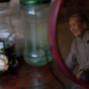 A still from The Apology. A round mirror sits atop a wooden surface. In the reflection is an older person, wearing a blue button-down shirt and facing to the side. Behind the mirror are two bottles.