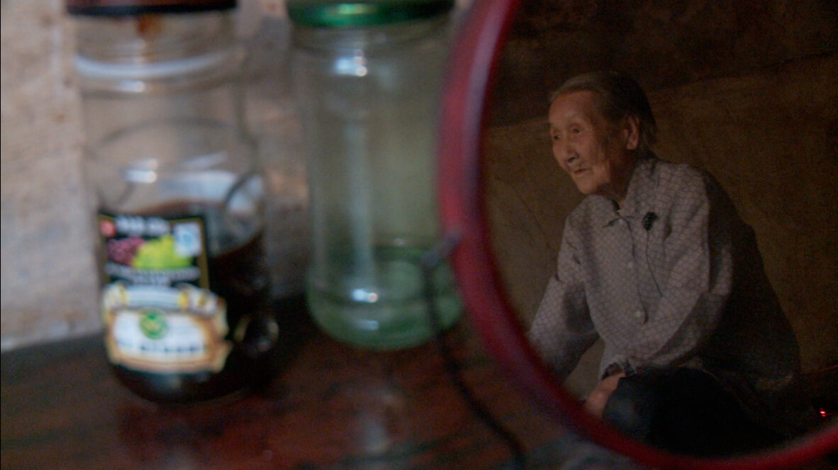 A still from The Apology. A round mirror sits atop a wooden surface. In the reflection is an older person, wearing a blue button-down shirt and facing to the side. Behind the mirror are two bottles.