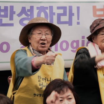 A still from The Apology. A person wearing a yellow smock with Korean text over the chest, a wide-brimmed straw hat, and glasses stands in front of a large banner also with Korean text. Their fist is raised pointing directly out in front of them.