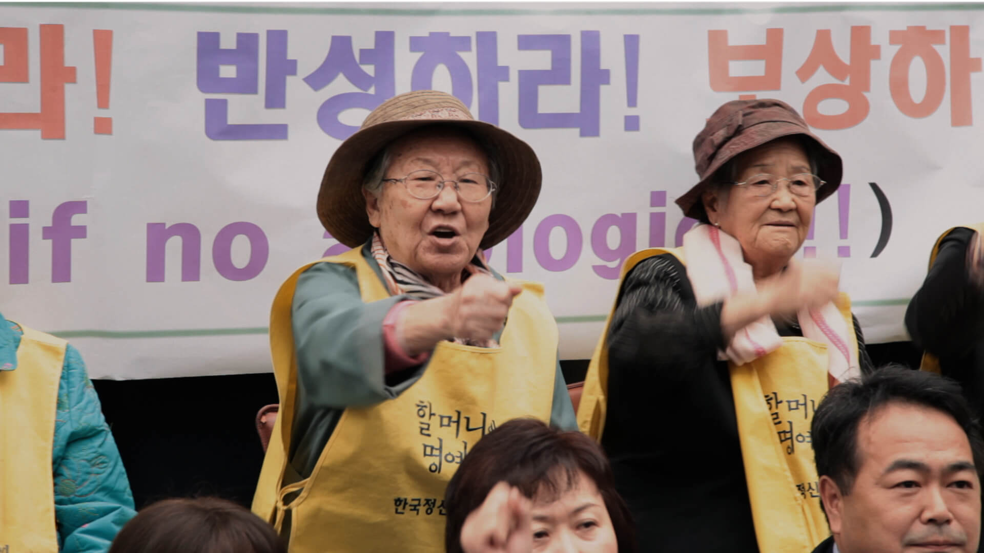 A still from The Apology. A person wearing a yellow smock with Korean text over the chest, a wide-brimmed straw hat, and glasses stands in front of a large banner also with Korean text. Their fist is raised pointing directly out in front of them.