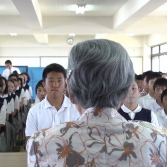 A still from The Apology. A person with grey hair has their back turned toward the camera. In front of them are four rows of children, seated at desks in a classroom and wearing uniforms.