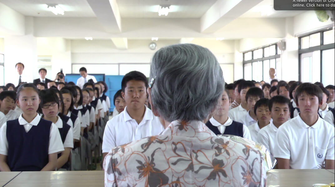 A still from The Apology. A person with grey hair has their back turned toward the camera. In front of them are four rows of children, seated at desks in a classroom and wearing uniforms.