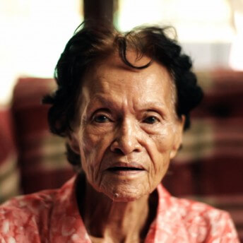 A photo of an elderly woman looking directly at the camera. She has black hair and is wearing a pink patterned shirt.