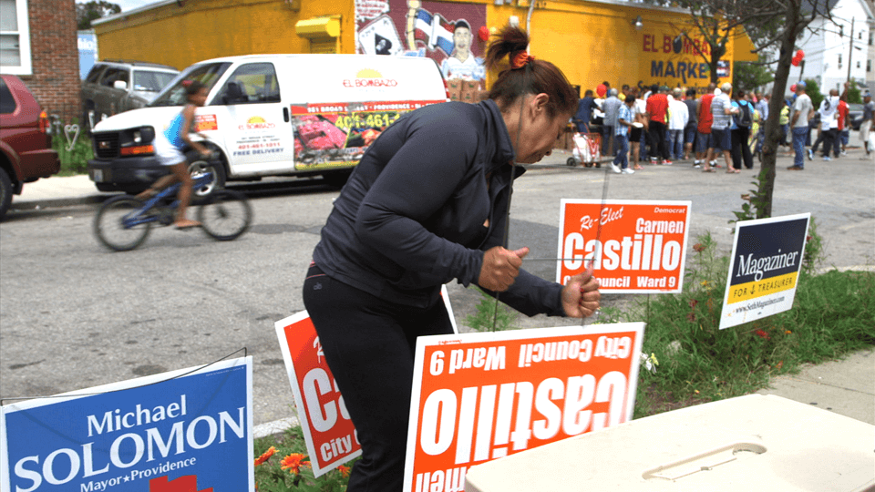 A woman placing posters on the grass that say "Re-elect Carmen Castillo"