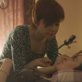 Still from Unrest. A young woman is putting eye makeup on another young woman lying in a bed.