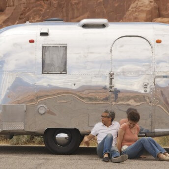 Still from Unrest. A man and a woman sitting on the ground in front of a small, silver airstream trailer. The background is the mountains of a red, sandy desert.